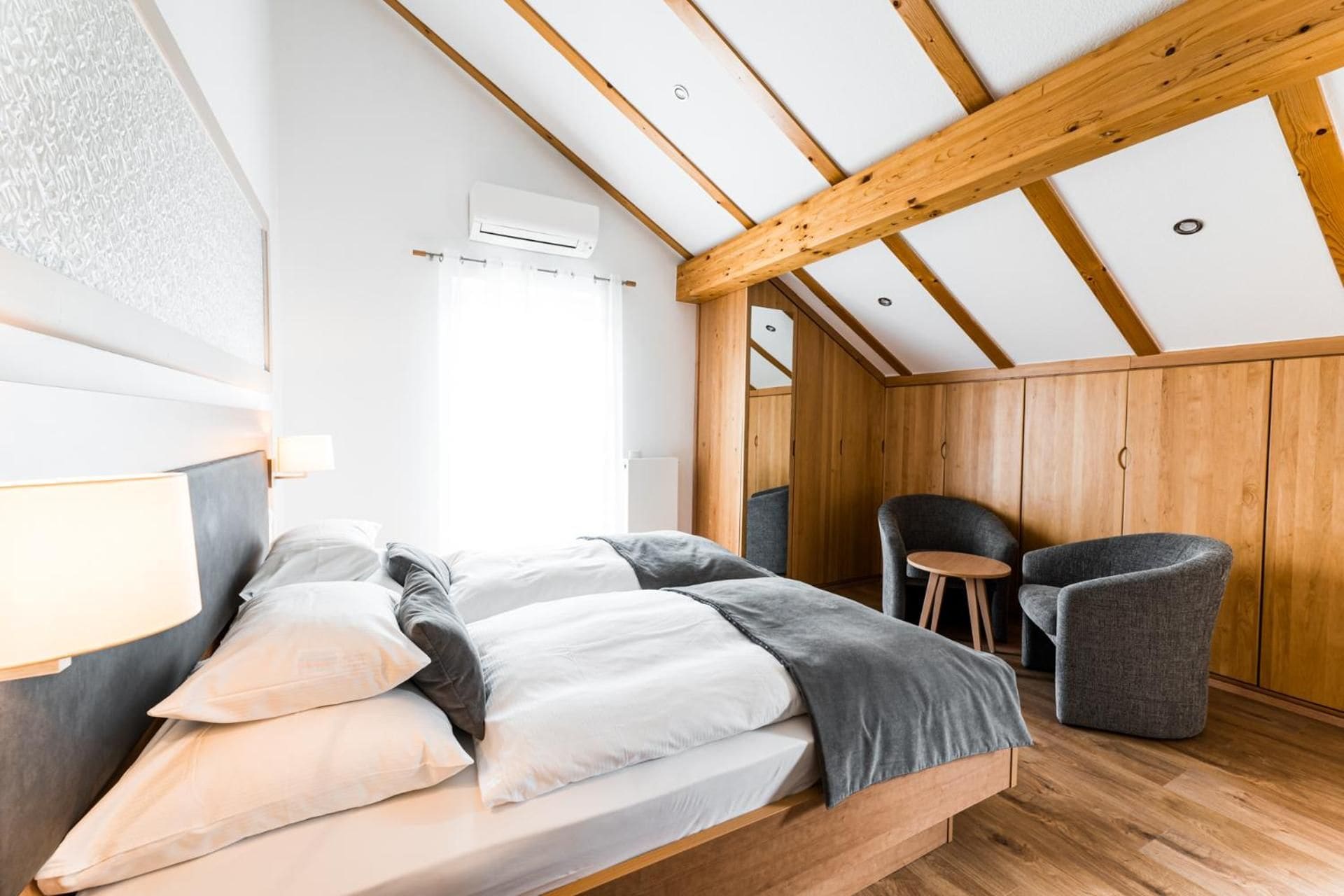 A bedroom with a sloped ceiling, wooden beams, a bed with pillows, a chair, and an air conditioning unit.