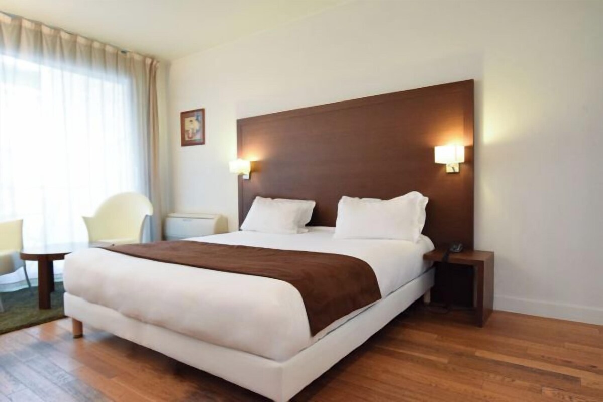 A hotel room with a large bed, two bedside tables, a chair, and a window with curtains.