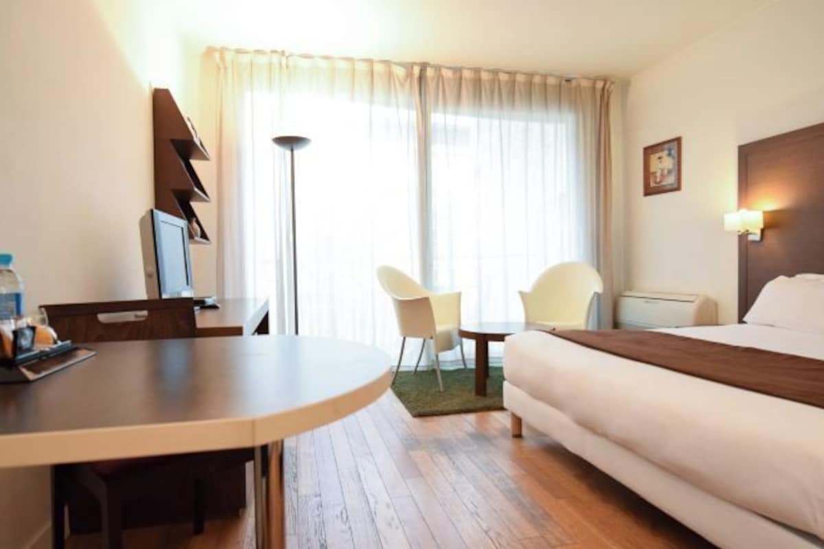 Una habitación de hotel con cama, escritorio, silla y un amplio ventanal con cortinas.
