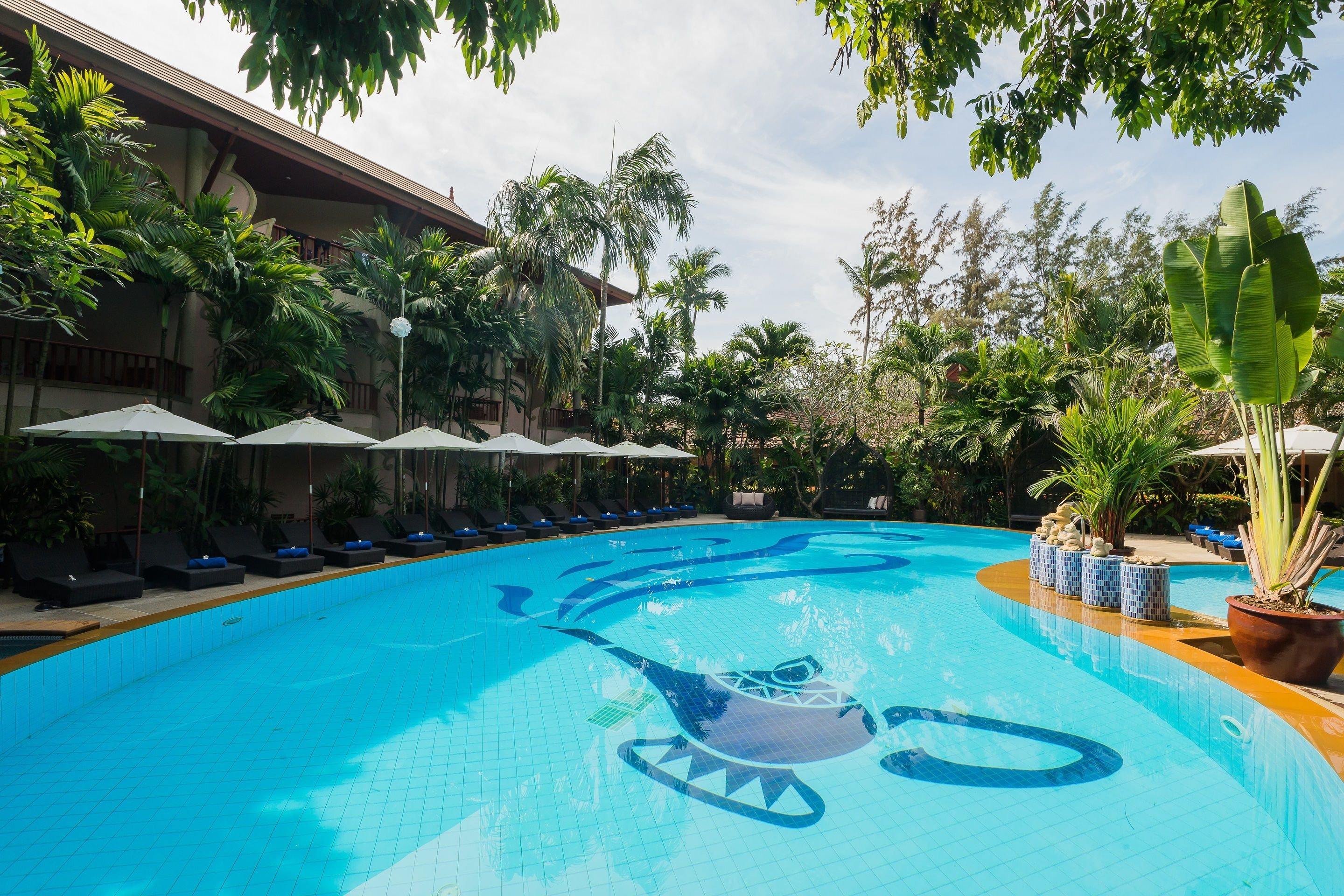 A swimming pool with a shark design, surrounded by lounge chairs and umbrellas, with palm trees and a building in the background.