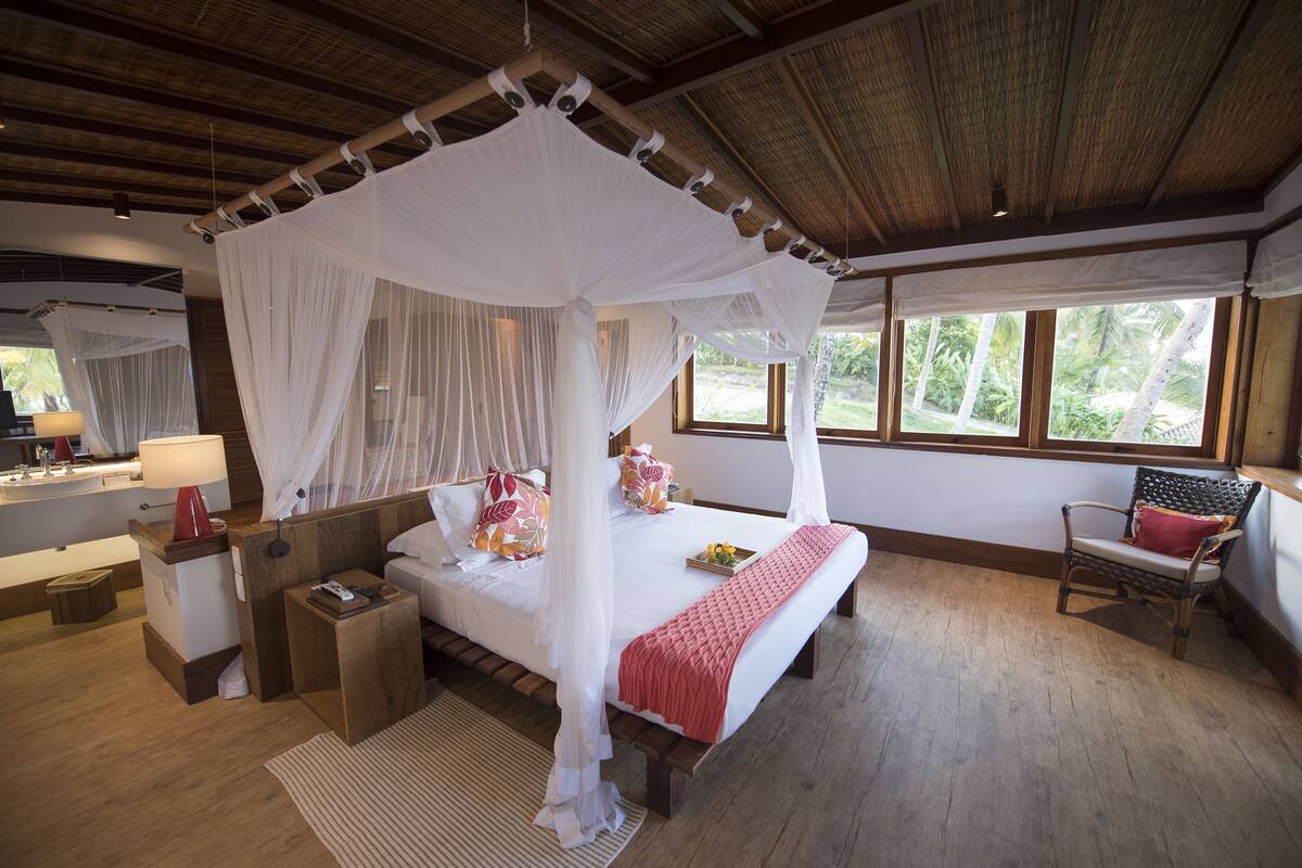 A bedroom with a four-poster bed, bedside tables, a chair, and a view of the outdoors.