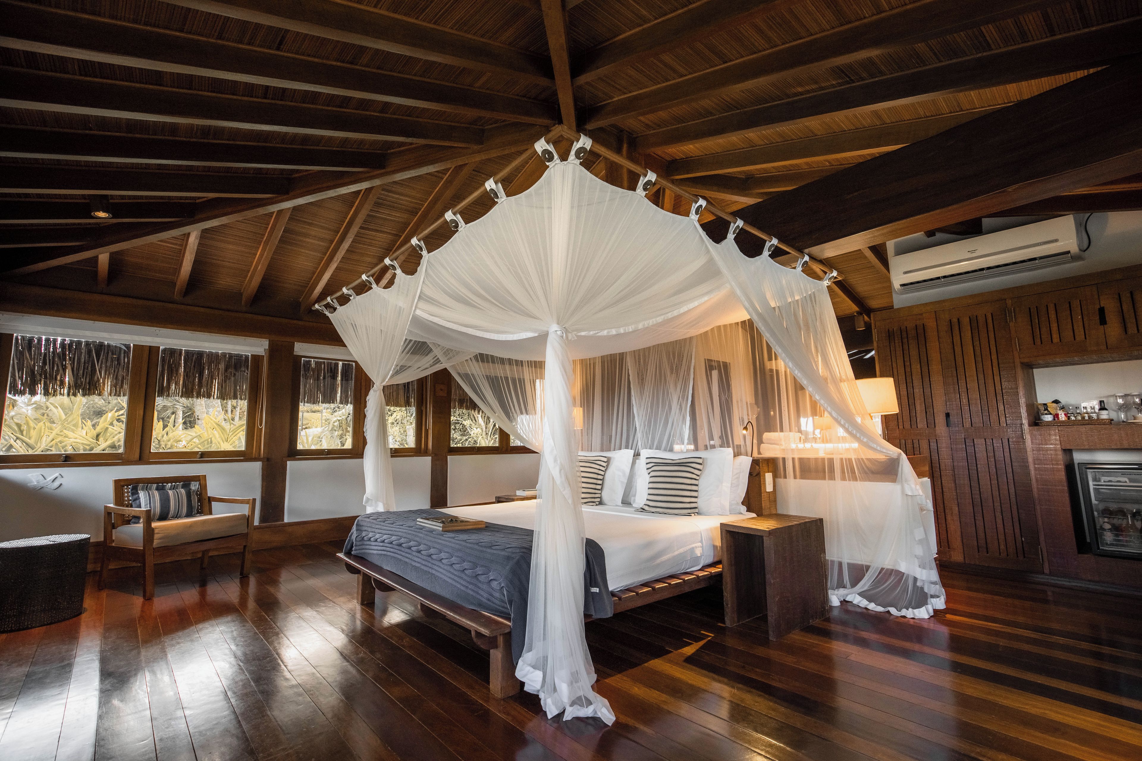 A room with a canopy bed, wooden flooring, a bench, and a view of the outdoors through large windows.