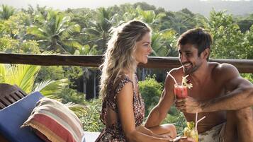 Un couple est assis sur une terrasse en bois, d’où l’on peut admirer une végétation luxuriante et des collines.