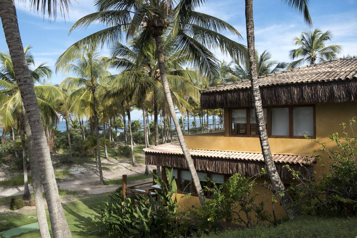 A yellow building with a thatched roof, surrounded by palm trees and a beach view.