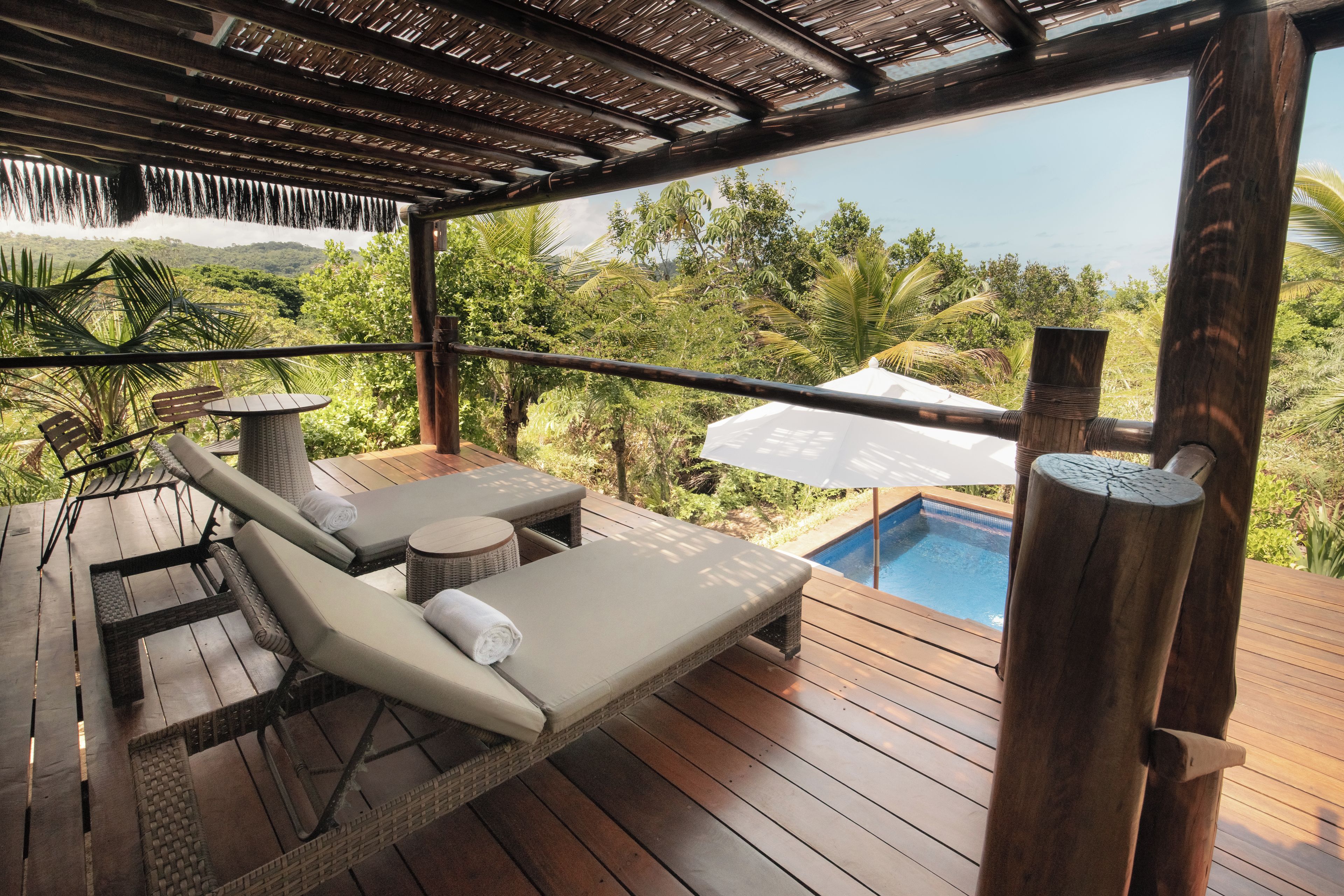 Terraza de madera con sillones de descanso y una mesita, con vistas a una piscina y exuberante vegetación.