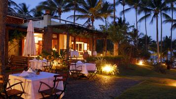 An outdoor dining area with tables and chairs, palm trees, and a building with a covered entrance.