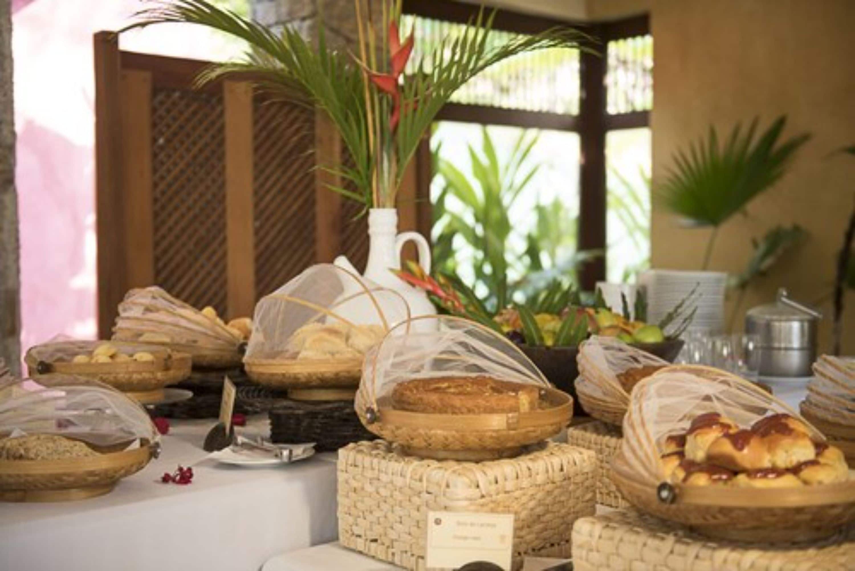 Uma mesa de buffet com diversos produtos de panificação em cestas, um jarro branco e uma pequena placa.