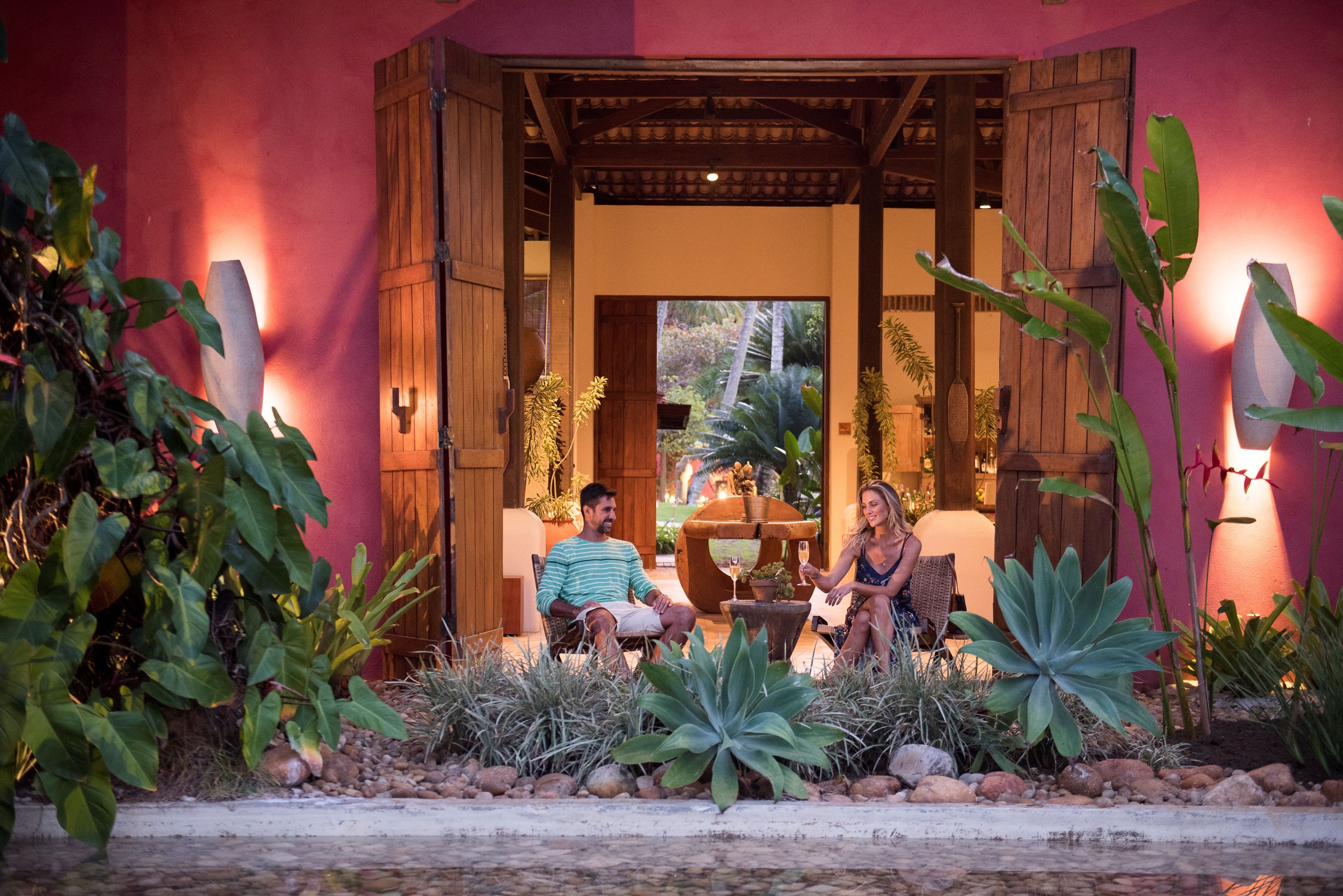Una pareja sentada en muebles de exterior en un jardín tropical con una pérgola de madera.