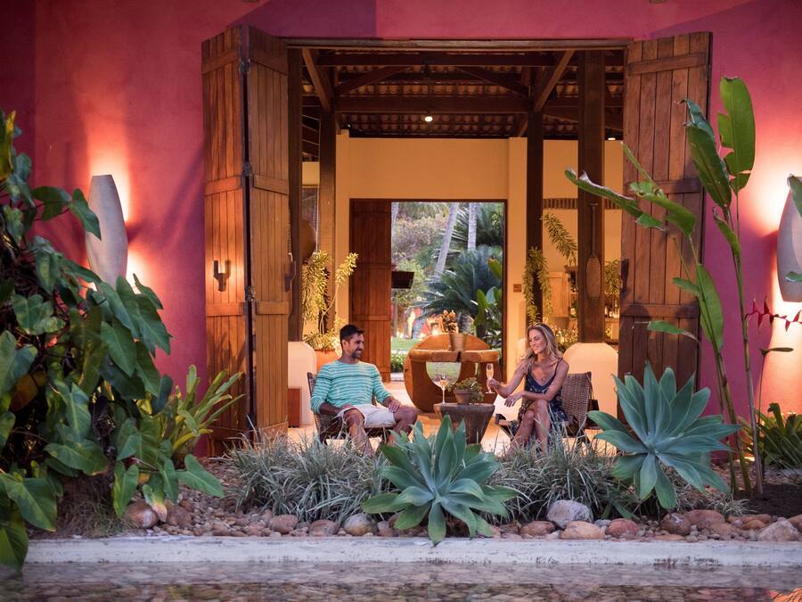 Una pareja sentada en muebles de exterior en un jardín tropical con una pérgola de madera.