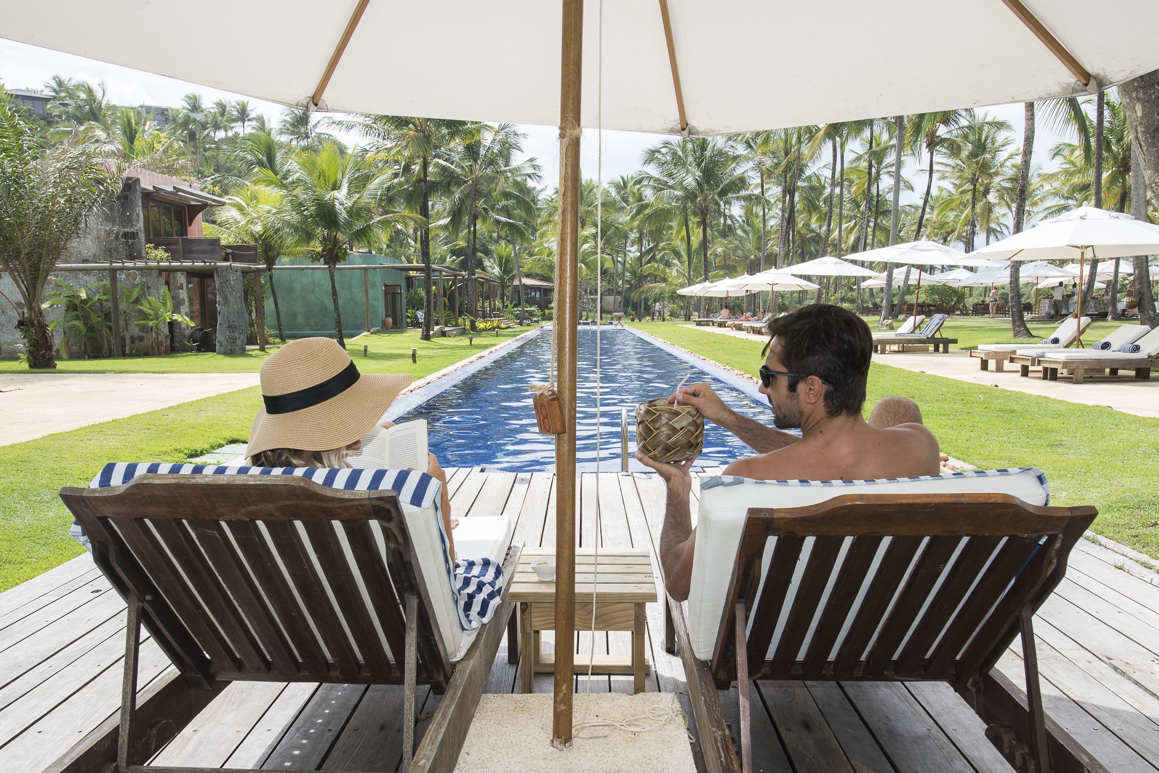 Un área junto a la piscina con sillones de madera, una piscina y palmeras al fondo.