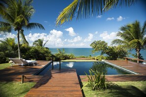 Una terraza de madera con piscina, palmeras y vista al océano.