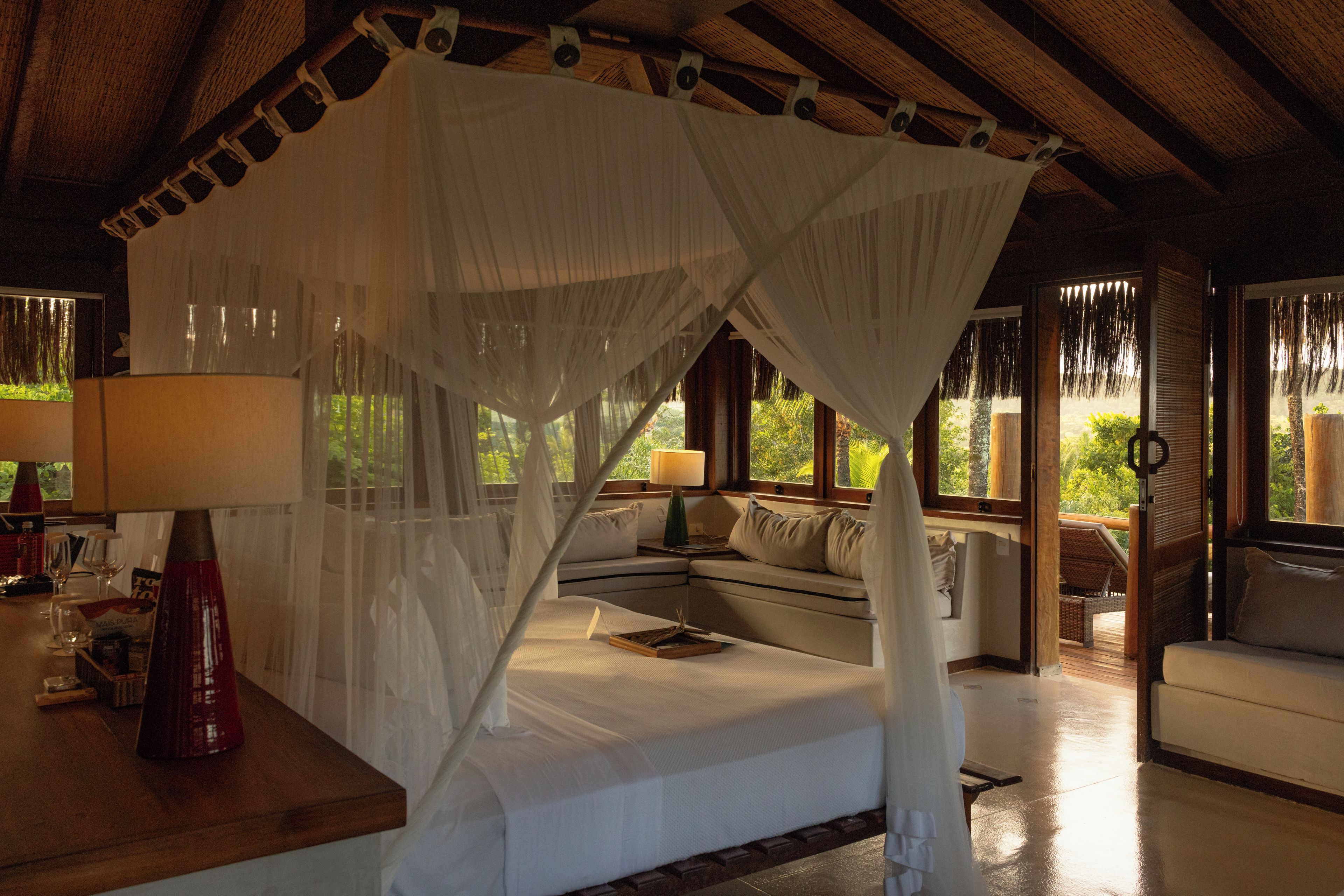 Une chambre avec un lit sous un dais, un canapé, une table avec une lampe et une vue sur la verdure à l’extérieur.