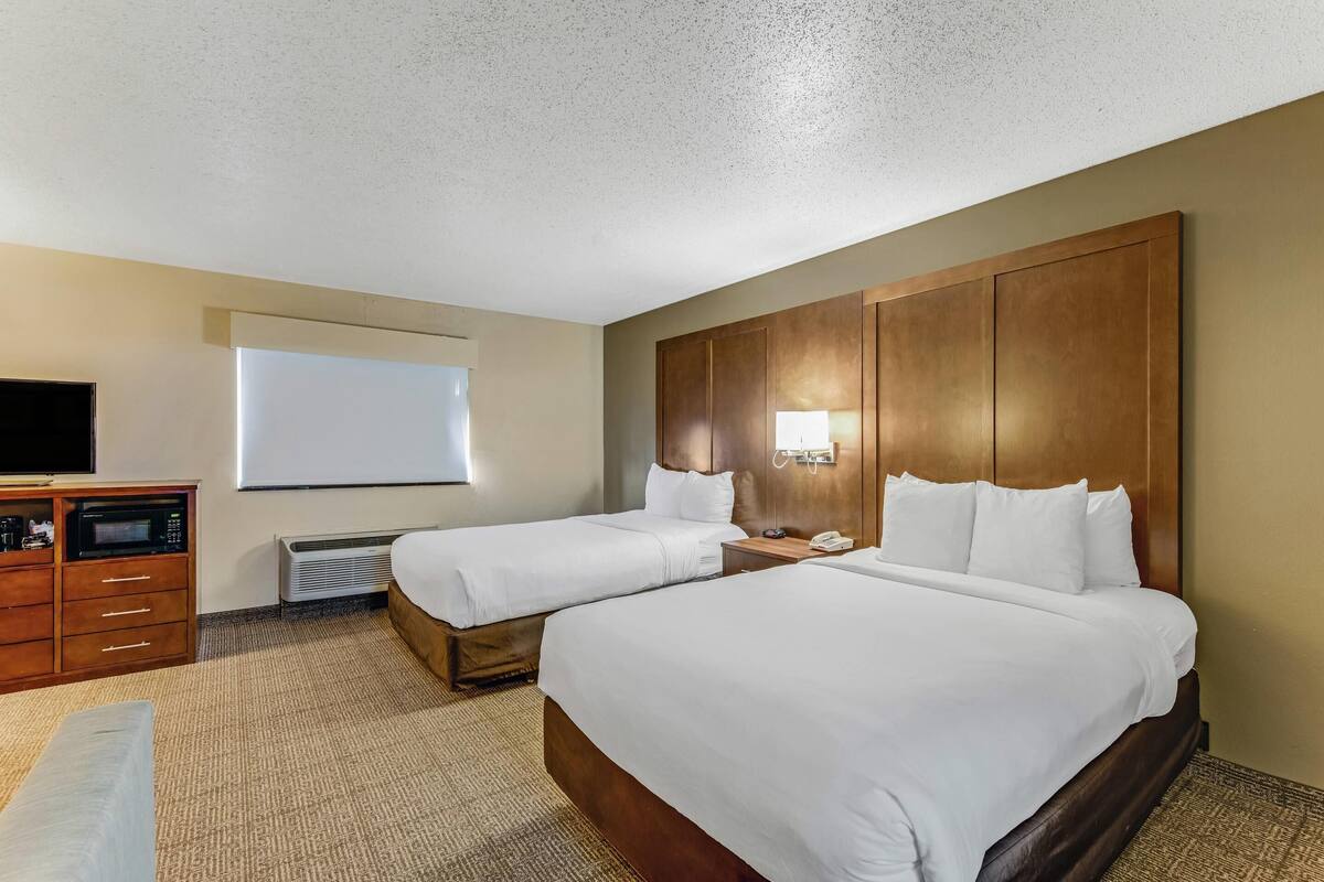 Une chambre d’hôtel avec deux lits, une tête de lit en bois, un téléviseur à écran plat, un bureau avec un four à micro-ondes et une fenêtre équipée d’un écran de projection.