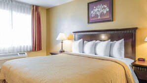 Quarto de hotel com uma cama grande, uma mesinha de cabeceira com abajur, um quadro na parede e uma janela com cortinas.