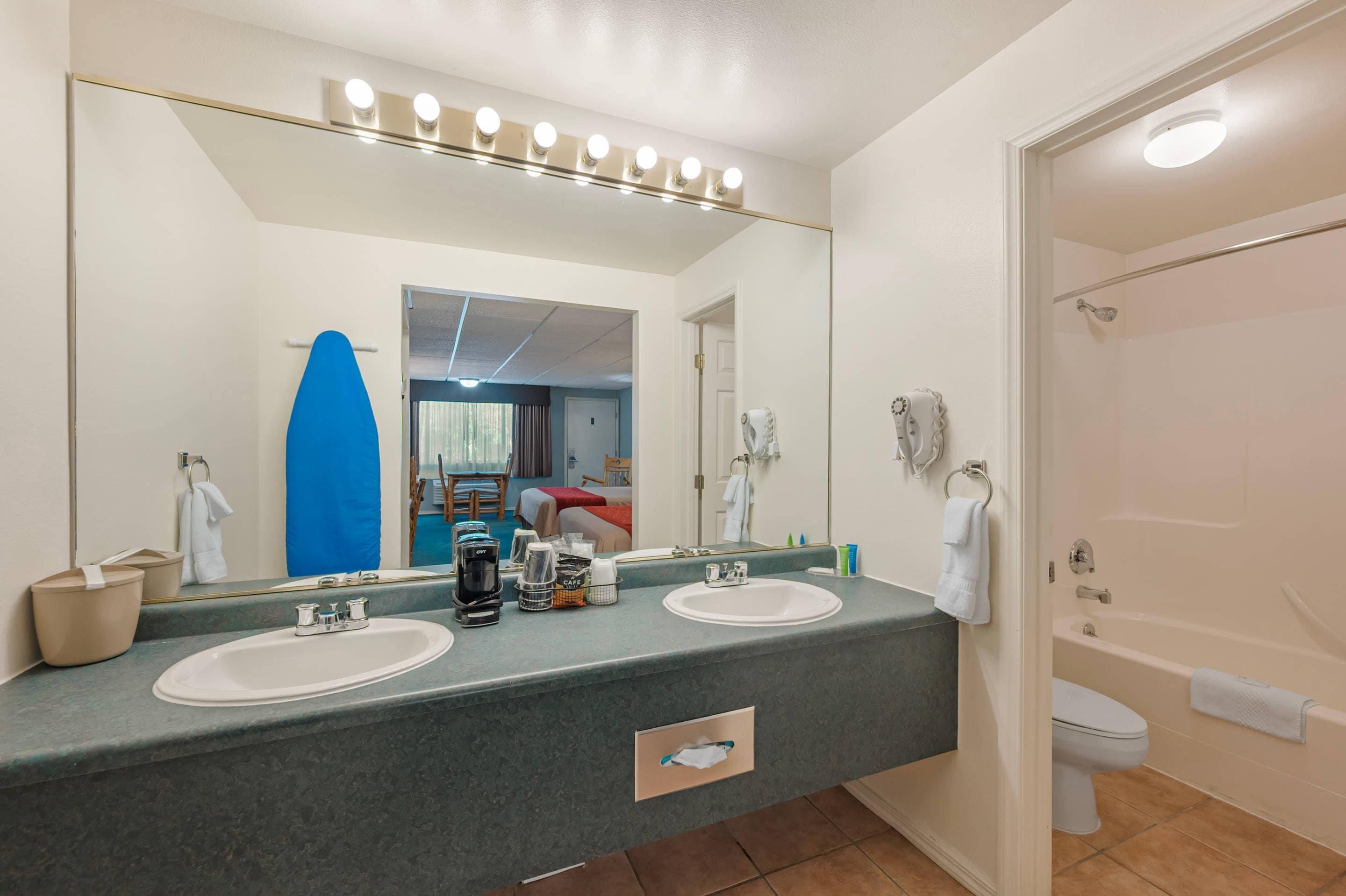 Une salle de bain avec deux lavabos, un grand miroir, une douche et une porte donnant sur une pièce où se trouvent un lit et une fenêtre.