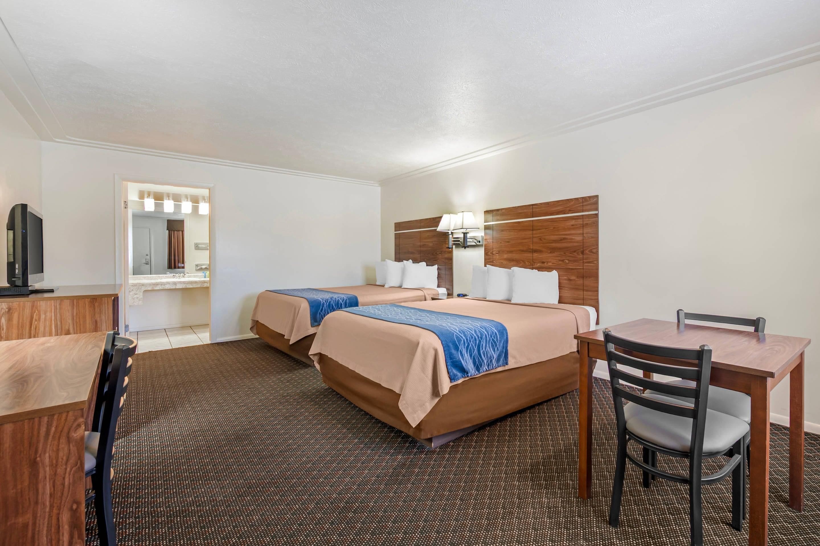 A hotel room with two beds, a desk, a chair, a television, and a bathroom visible through an open door.