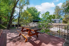 Une terrasse en bois, agrémentée de tables et de chaises pour pique-niquer, entourée d’arbres et d’un mur de pierres.