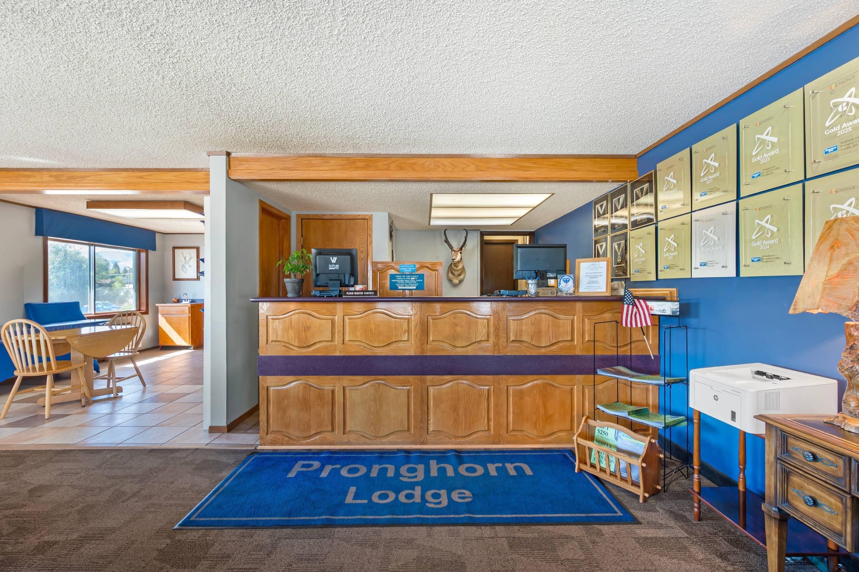 Vastaanottoalue, jossa on puinen työpöytä, sininen matto, jossa on teksti ”Pronghorn Lodge”, seinä, jossa on kehystettyjä palkintoja, ja näkymä huoneeseen, jossa on sänky ja ikkuna.