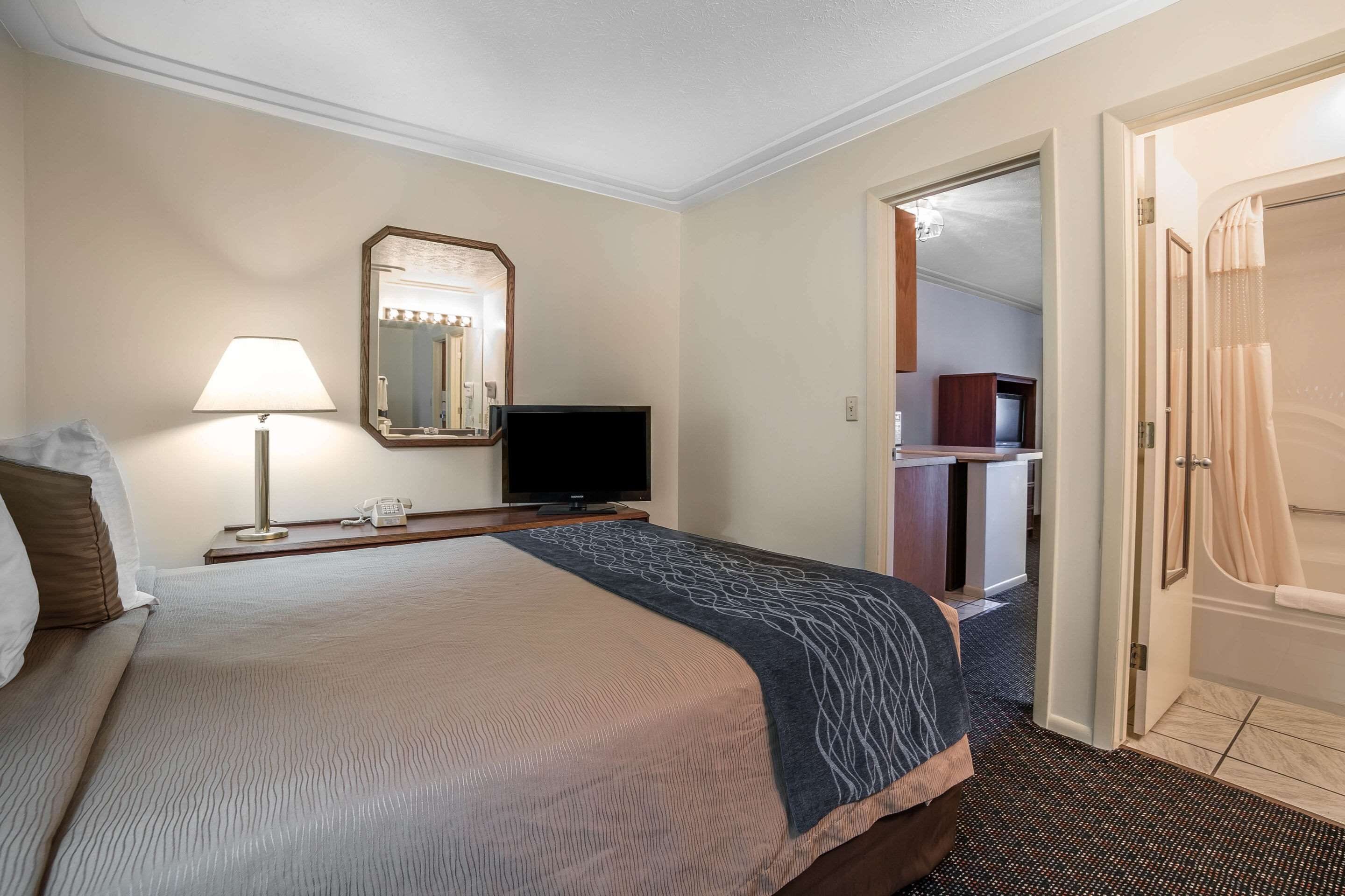 Una habitación de hotel con una cama, televisión, lámpara, espejo y puerta al baño.