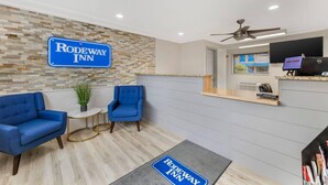 A reception area with a sign reading "Rodeway Inn," a reception desk, a seating area with blue chairs, a small table with a plant, and a welcome screen. - Rodeway Inn Lyons - La Grange (Lyons)