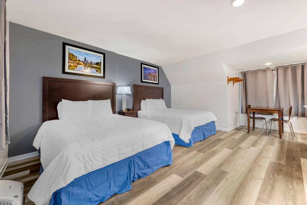 Quarto de hotel com duas camas, piso de madeira, uma escrivaninha com cadeiras e dois quadros na parede.