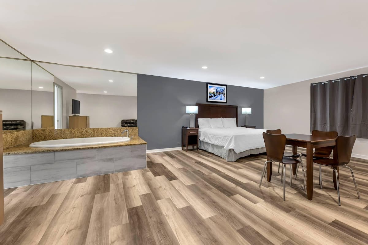 Quarto de hotel moderno com uma cama grande, área de estar com mesa e cadeiras, banheira integrada e um espelho grande.