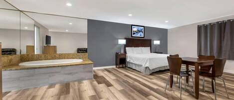 A modern hotel room with a large bed, a sitting area with a table and chairs, a built-in bathtub, and a large mirror.
