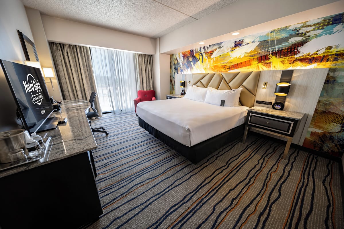 Habitación de hotel con una cama grande, un escritorio con computadora, una silla, un televisor y un ventanal con cortinas.