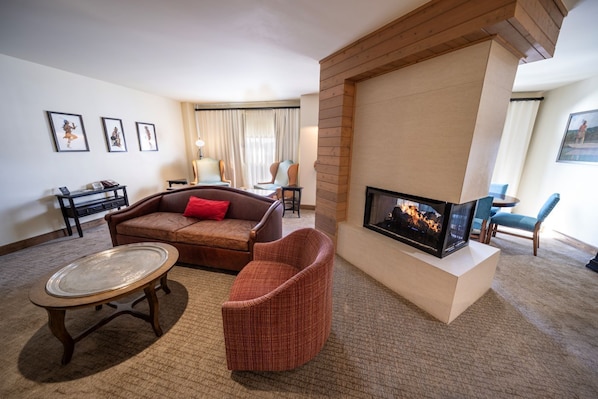 Una sala de estar con sofá, sillón y chimenea. Hay cuadros en la pared y una mesita.