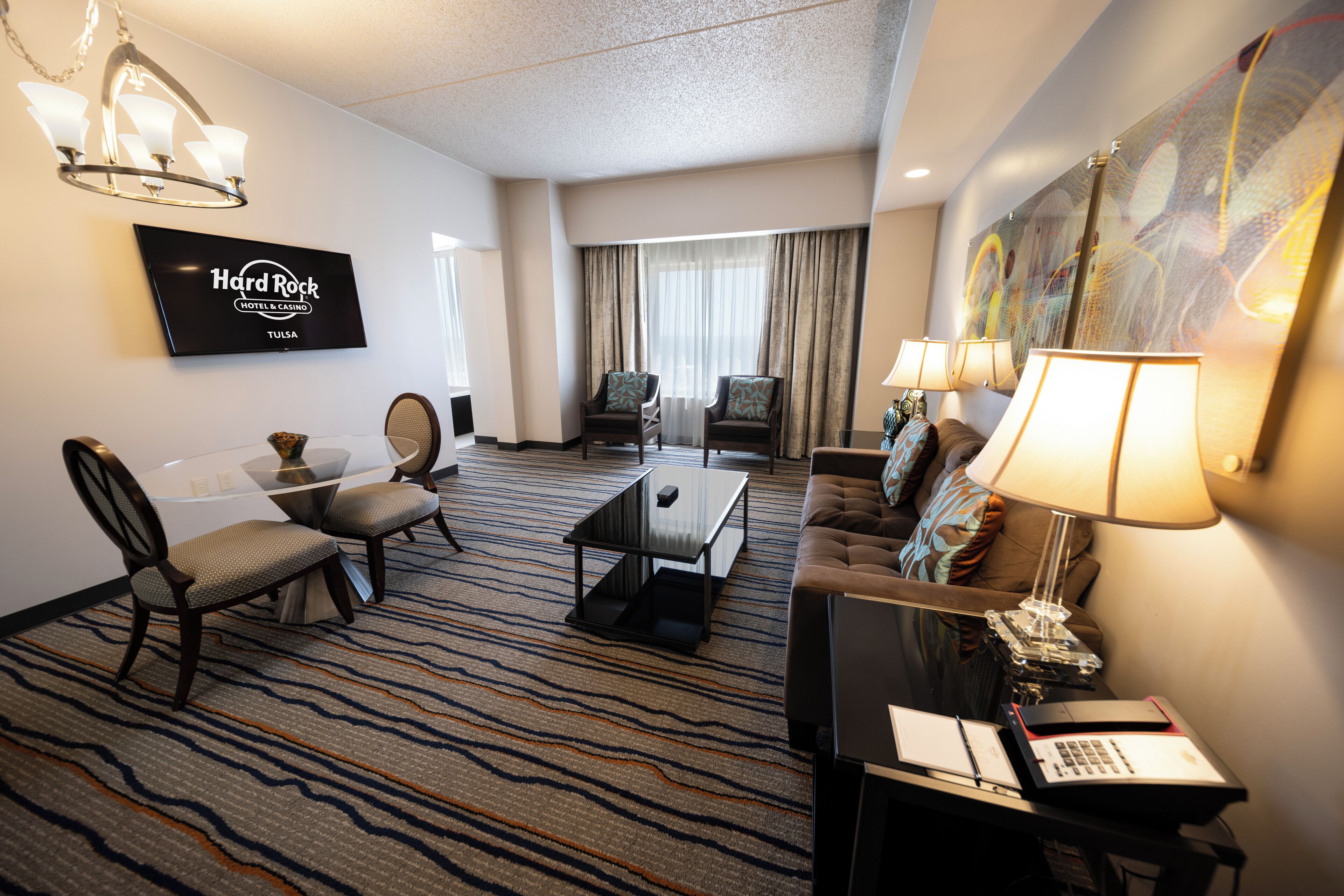 A hotel room with a flat-screen TV, a dining area with a glass table, a sofa, and a desk with a lamp.