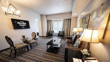 A hotel room with a flat-screen TV, a dining area with a glass table, a sofa, and a desk with a lamp.