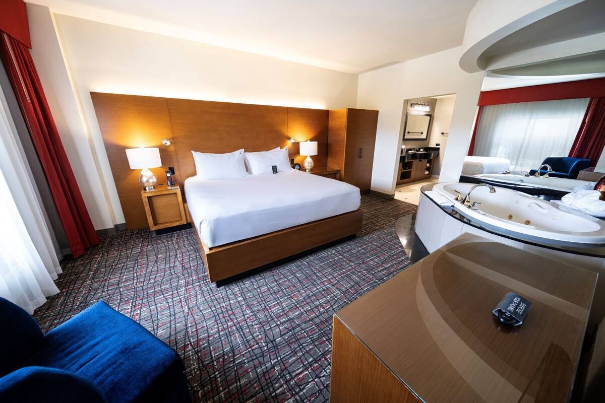 Habitación de hotel con una cama grande, lámparas de noche, un escritorio con silla, un baño con lavamanos y bañera, y un espejo.