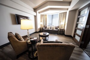 Habitación de hotel con sofá, sillones, una mesa de centro, televisor y un amplio ventanal con cortinas.