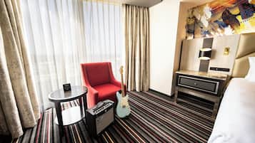 A hotel room with a red armchair, a guitar, a small table with a lamp, and a bed.
