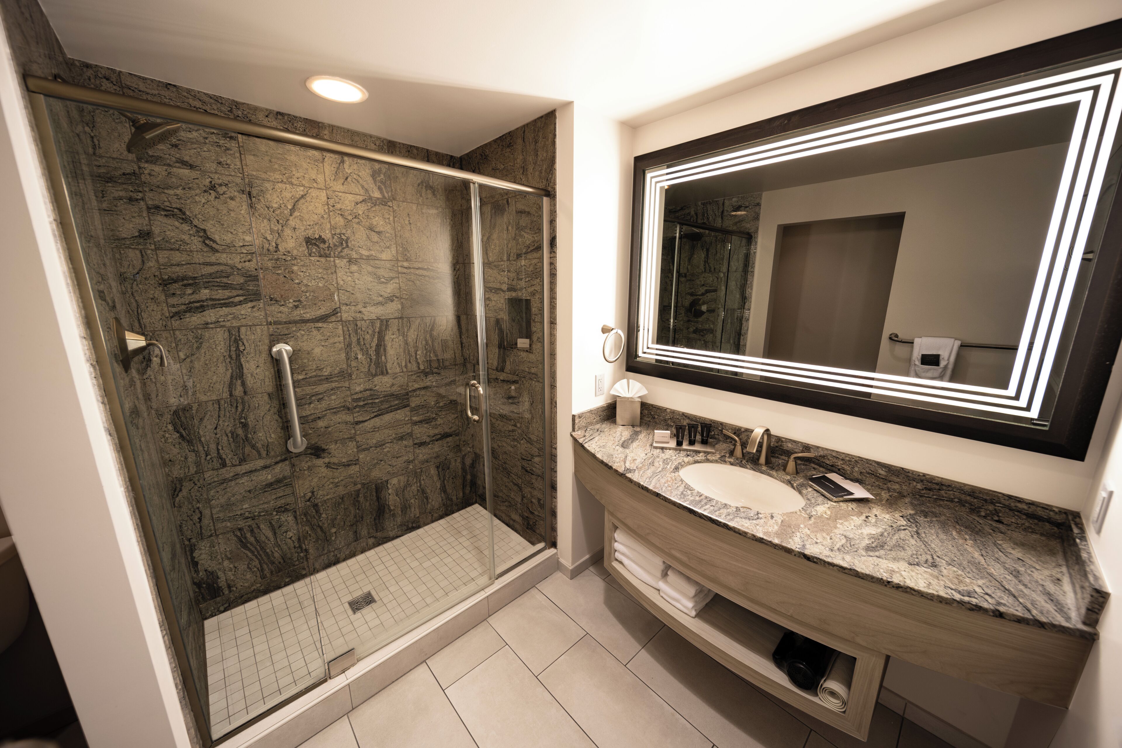 A modern bathroom with a glass-enclosed shower, marble countertops, and a large mirror with LED lighting.