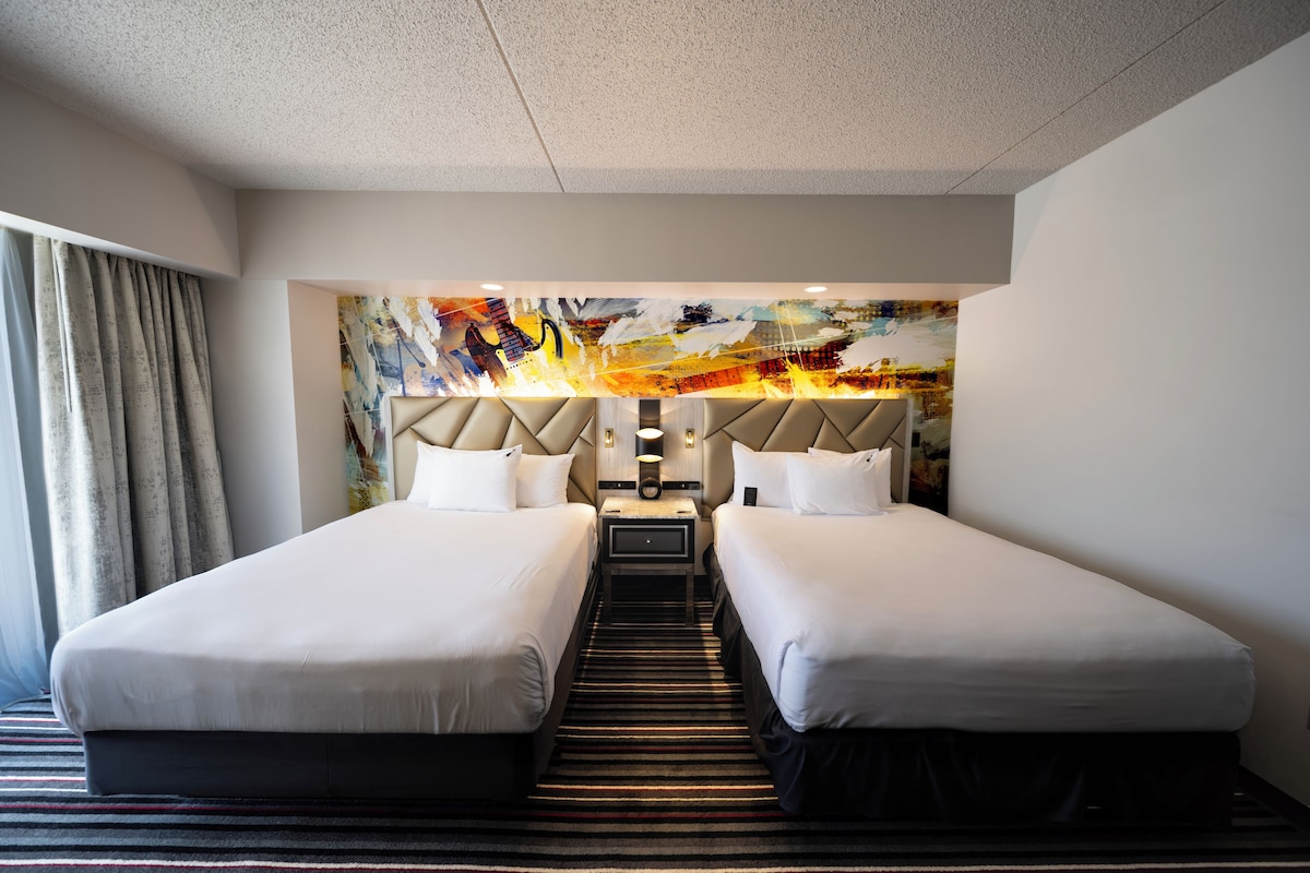 Habitación de hotel con dos camas, alfombra a rayas, cabecera moderna y un cuadro abstracto colorido en la pared.