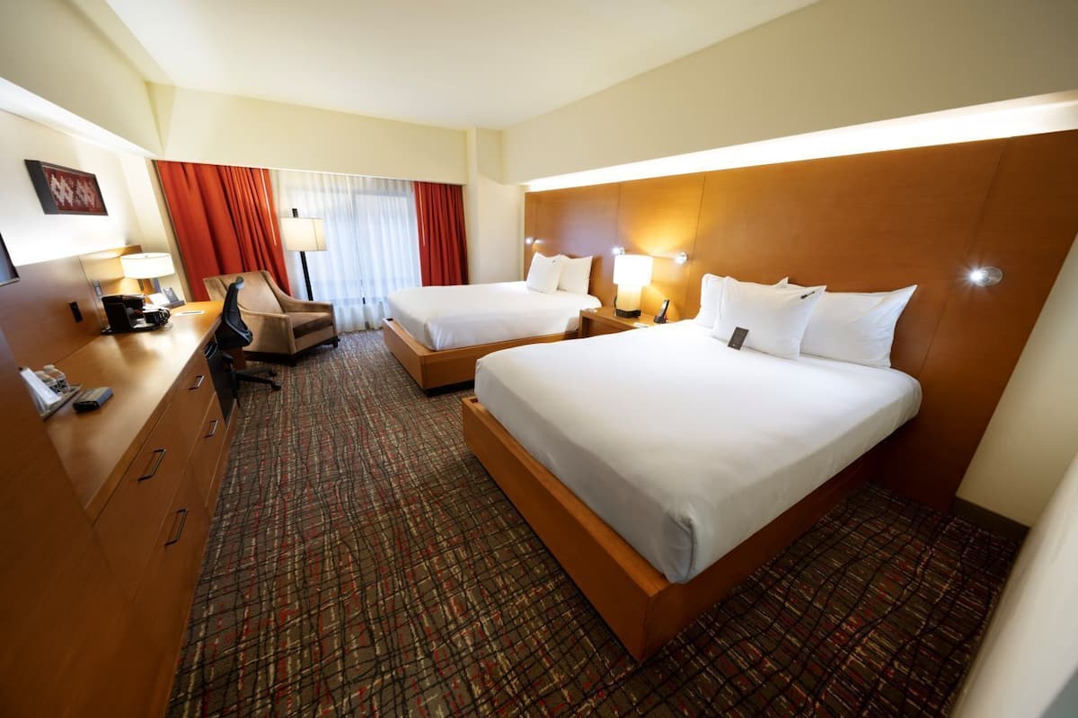 Una habitación de hotel con dos camas, un escritorio con silla, una lámpara y un ventanal con cortinas.