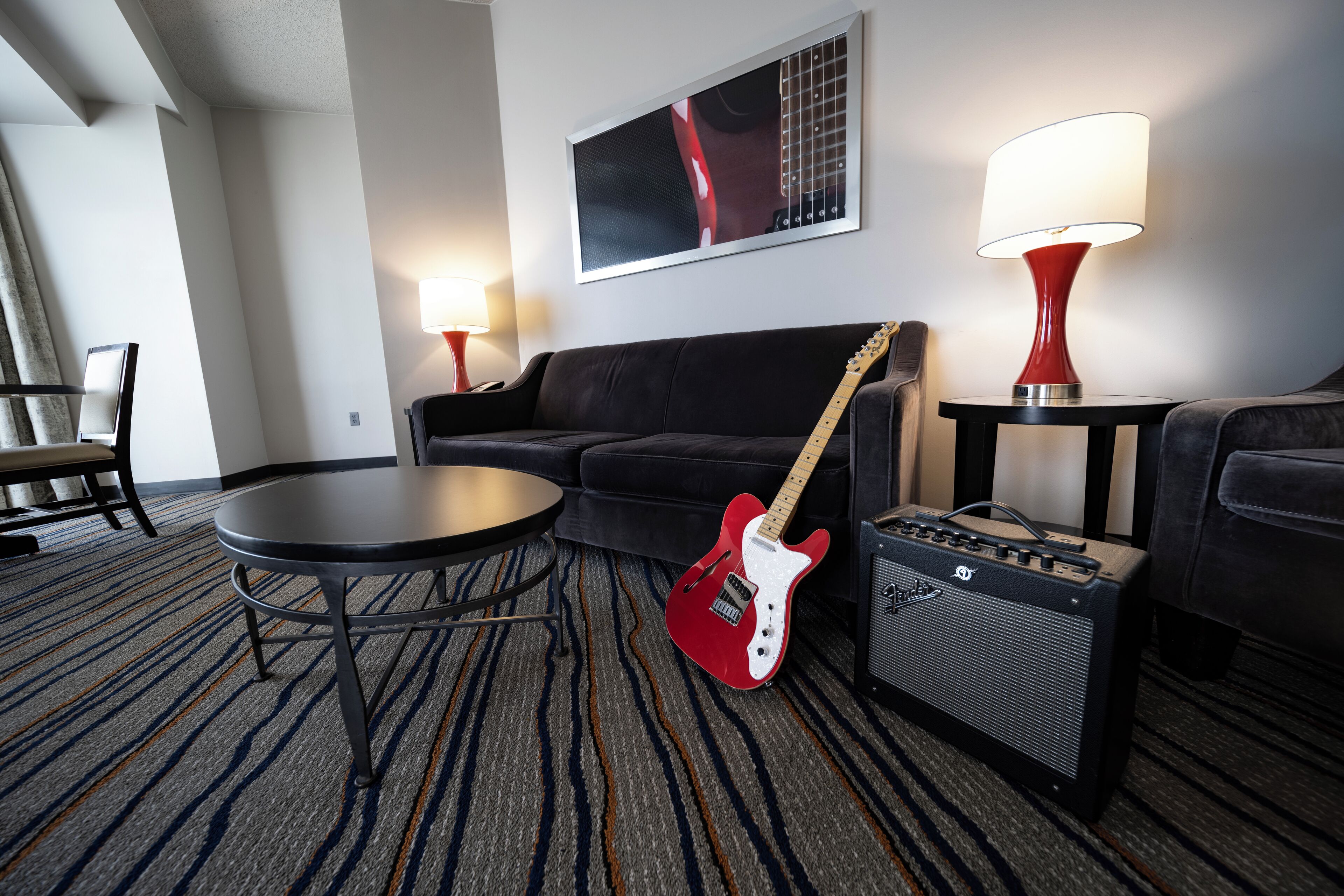 A hotel room with a black sofa, a small round table, a red electric guitar, a black amplifier, and two wall-mounted lamps.