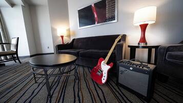 A hotel room with a black sofa, a small round table, a red electric guitar, a black amplifier, and two wall-mounted lamps.