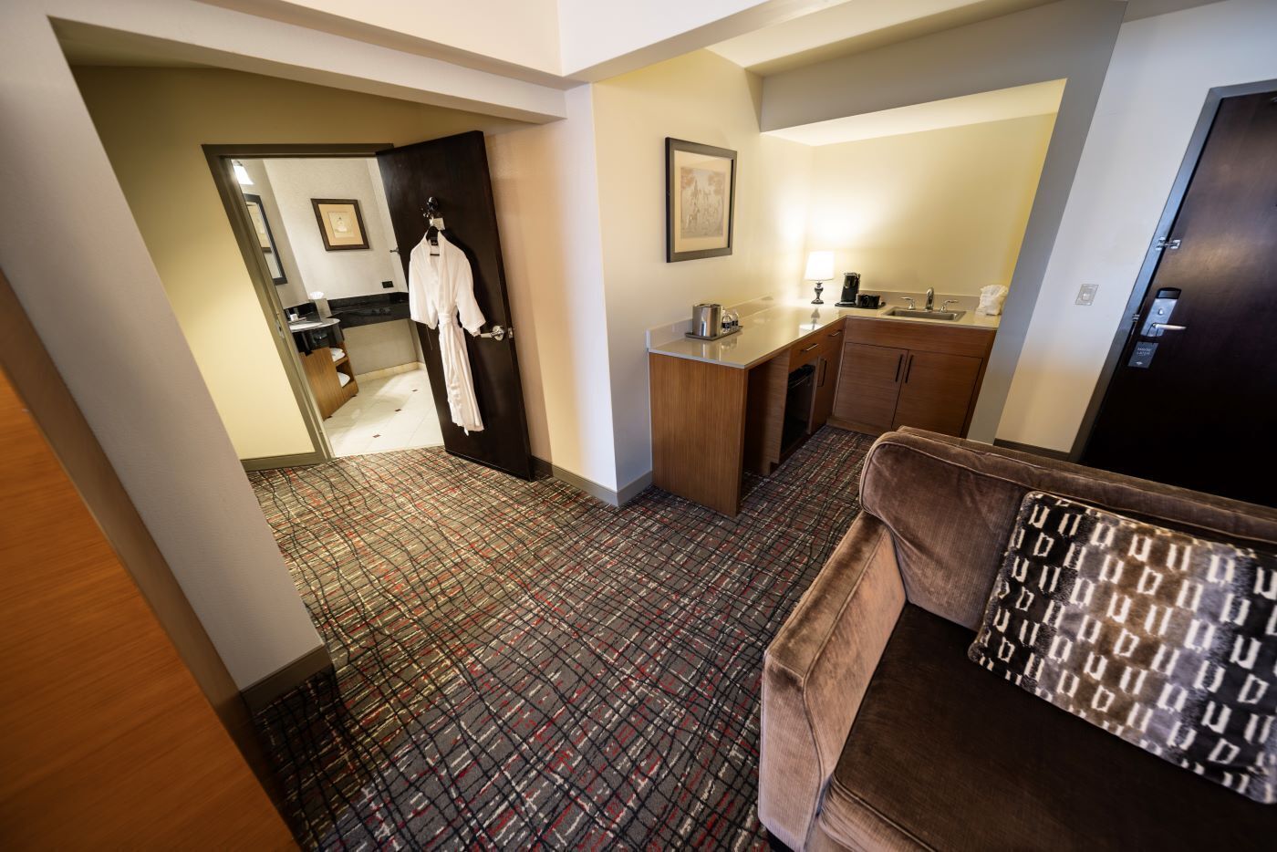 Habitación de hotel con alfombra estampada, un sofá marrón, una kitchenette pequeña y un baño visible a través de una puerta abierta.