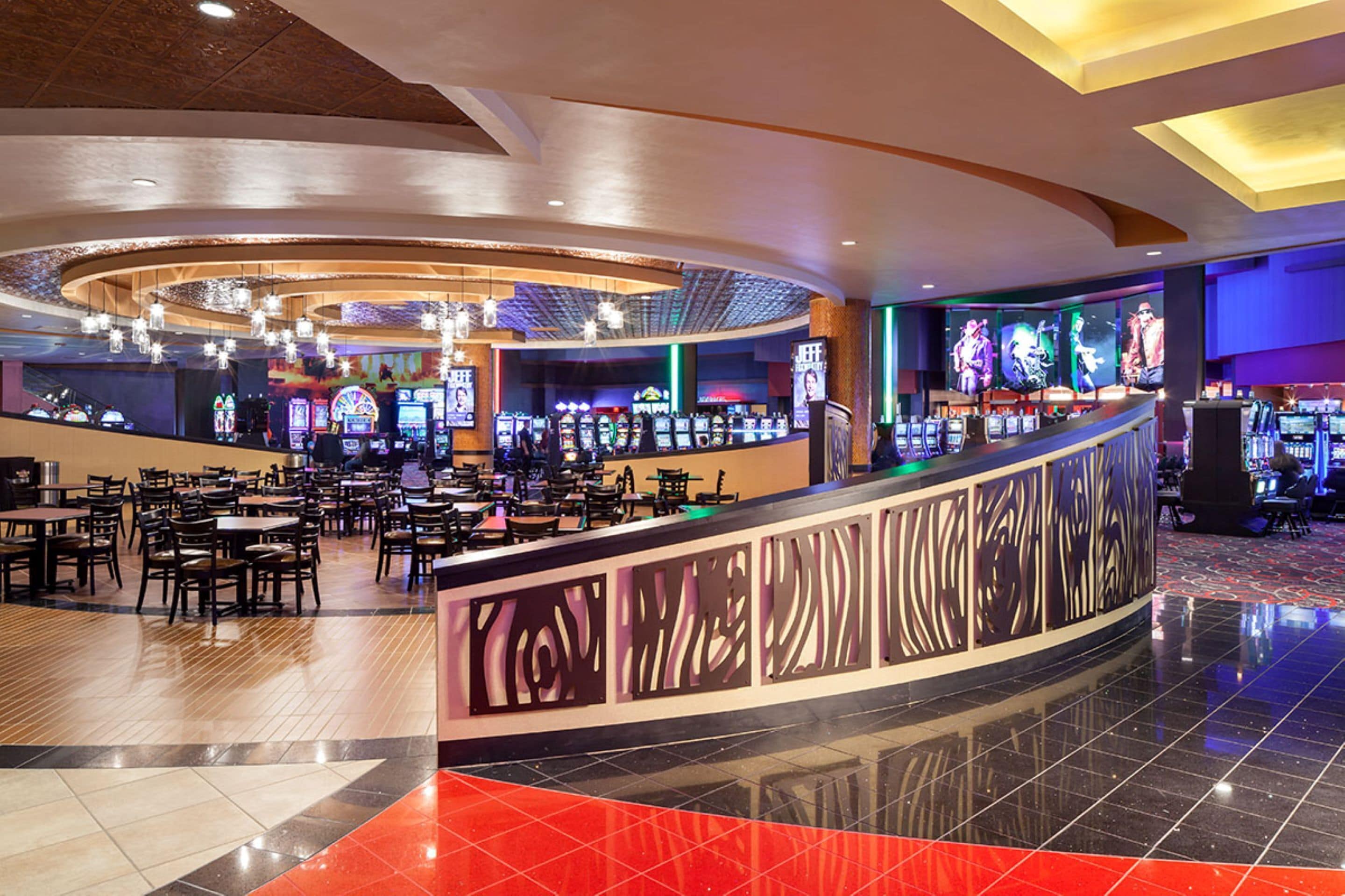 A casino with multiple gaming tables and chairs, a curved bar area, and a variety of slot machines.
