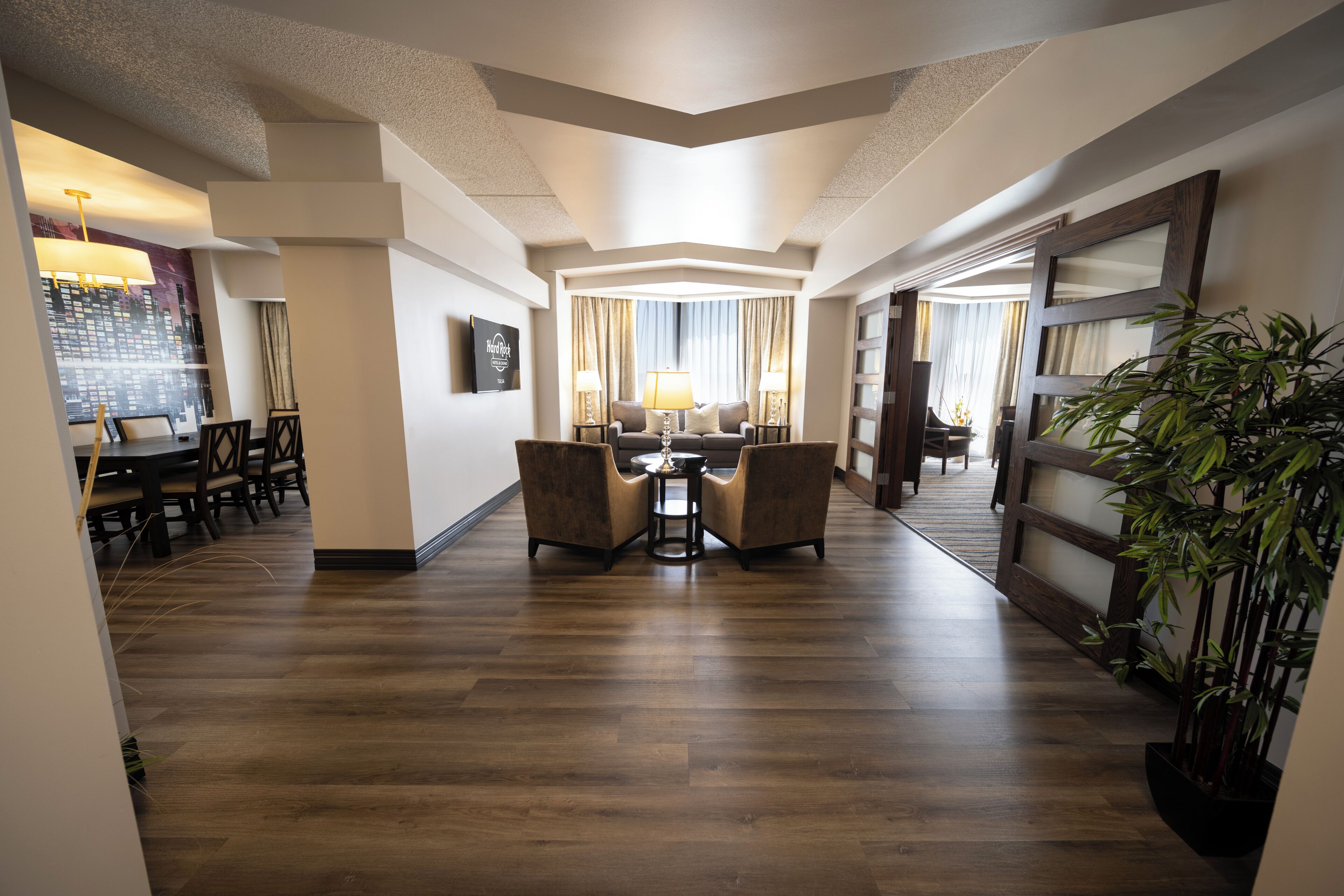 A spacious hotel lobby with a wooden floor, a central seating area with a table and chairs, and a large potted plant.