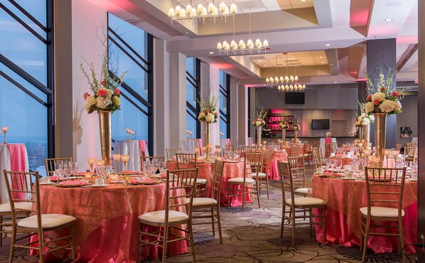 Un salón de banquetes con mesas redondas cubiertas con manteles rojos, sillas doradas y altos arreglos florales.