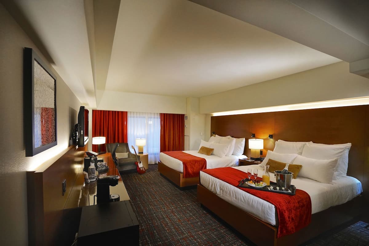Habitación de hotel con dos camas, un escritorio con computadora, una silla, una guitarra y una ventana con cortinas.