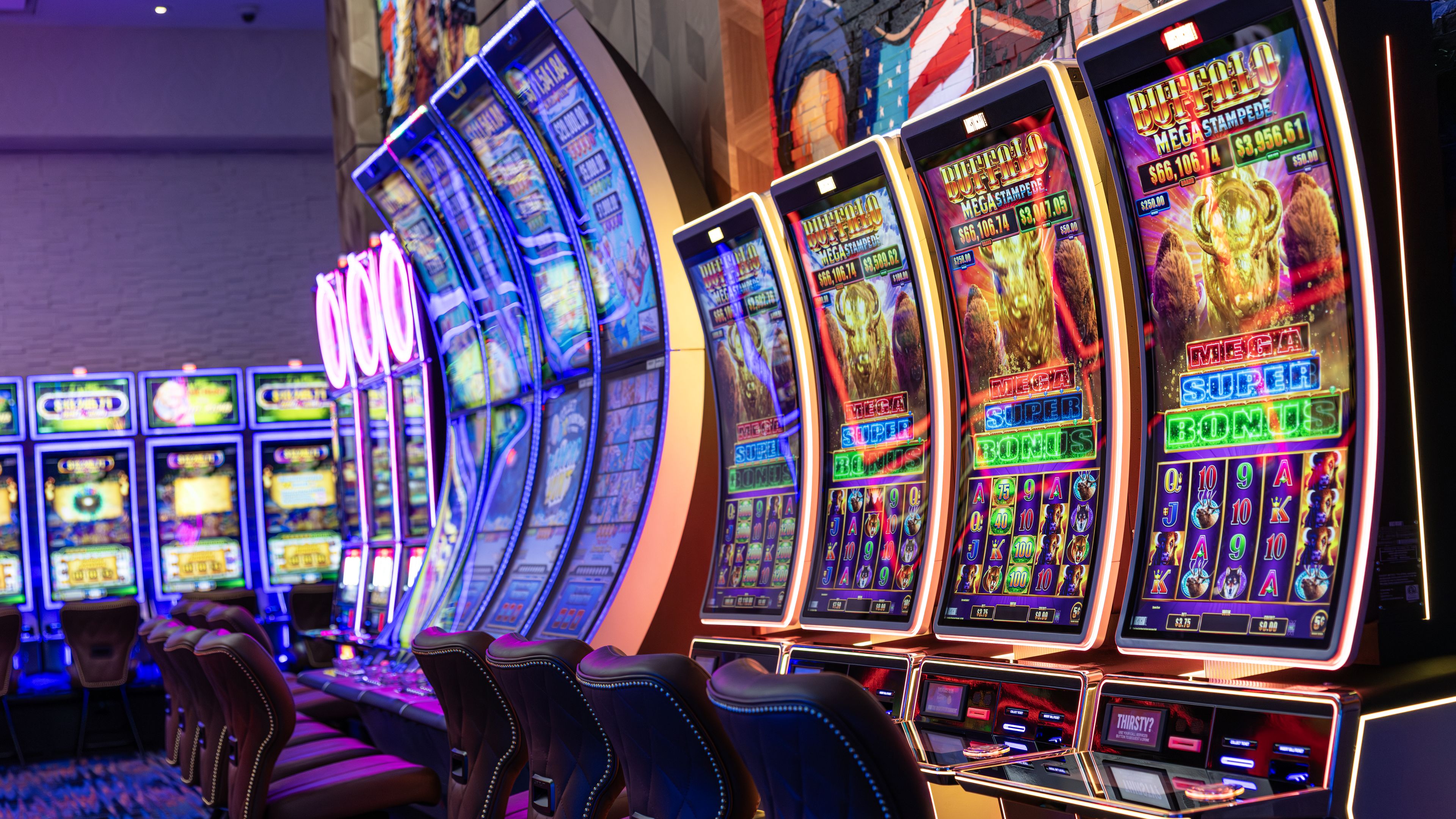 A row of slot machines with illuminated screens and a row of leather chairs in front.