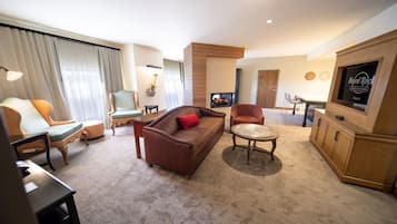 A hotel room with a brown sofa, a green armchair, a wooden coffee table, a TV, and a fireplace.