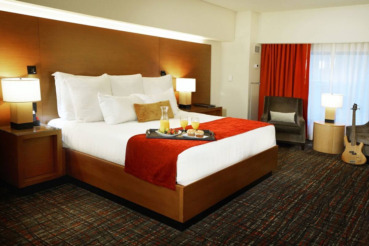 Habitación de hotel con una cama grande, dos mesitas de noche, una silla, una guitarra y una bandeja de desayuno sobre la cama.