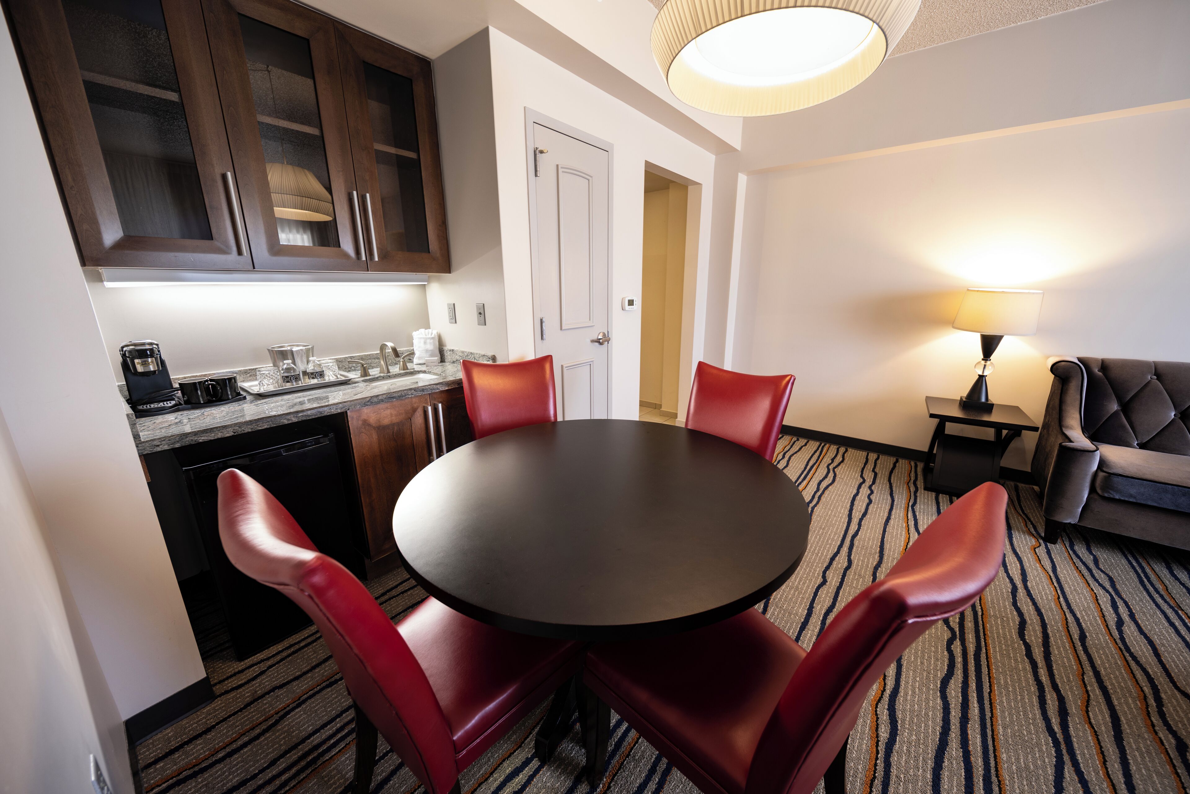 A hotel room with a dining area featuring a round table and red chairs, a kitchen with dark wooden cabinets, a sofa, and a lamp.