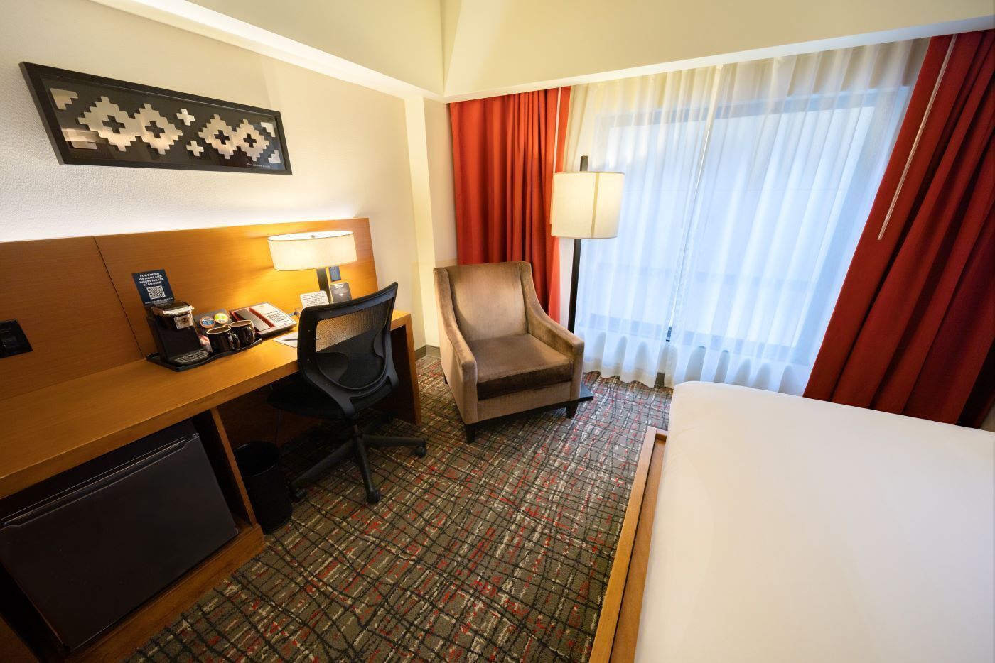 A hotel room with a bed, a desk with a computer, a chair, a coffee maker, and a window with curtains.