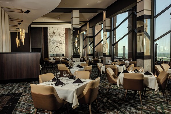 Un restaurante moderno con techos altos, ventanas amplias y una pared de mármol. Las mesas están dispuestas con manteles blancos y sillas marrones.